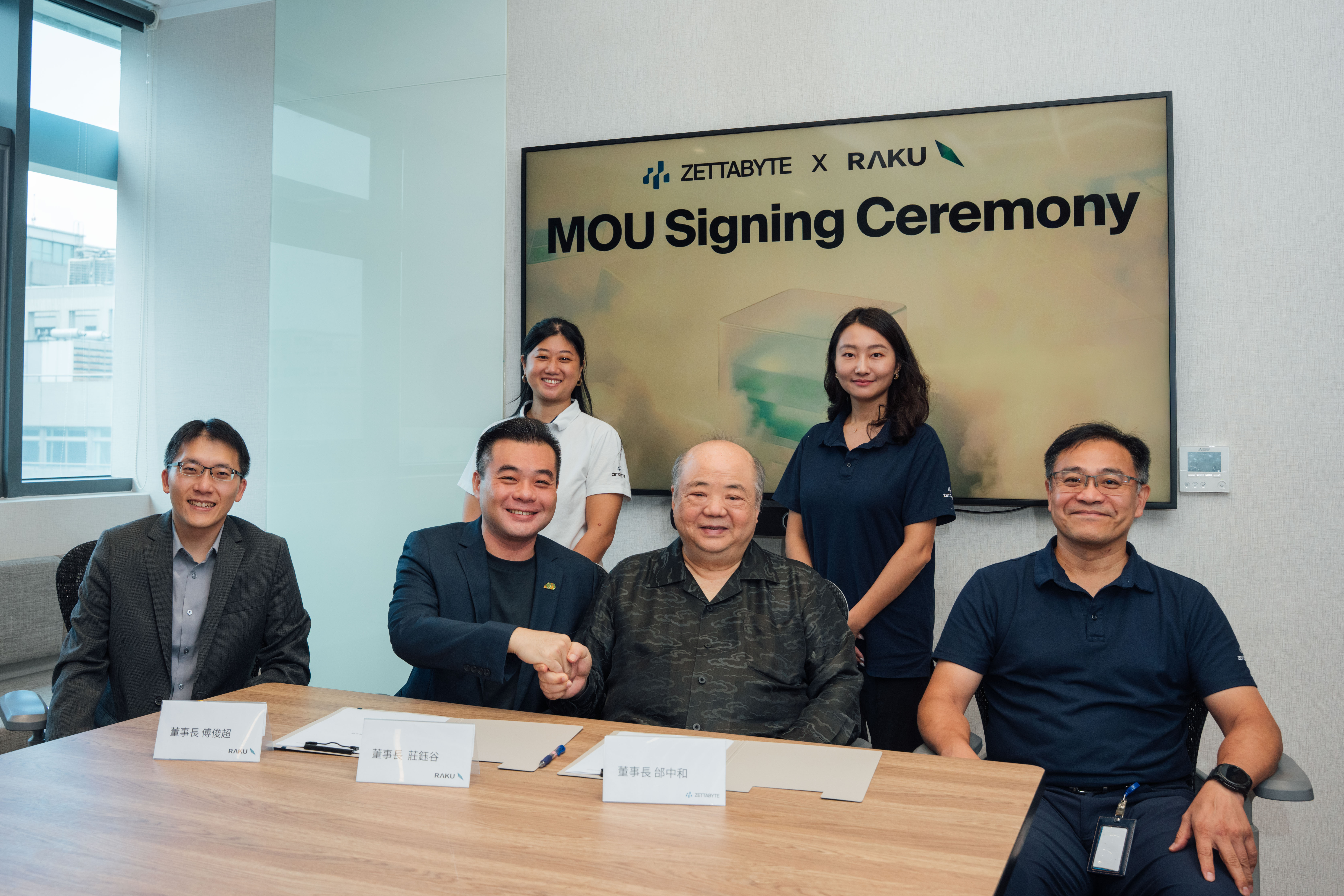 Representatives of Zettabyte and Raku Advanced Tech shaking hands across a signing table, with a screen behind them reading MOU Signing Ceremony under both company logos.